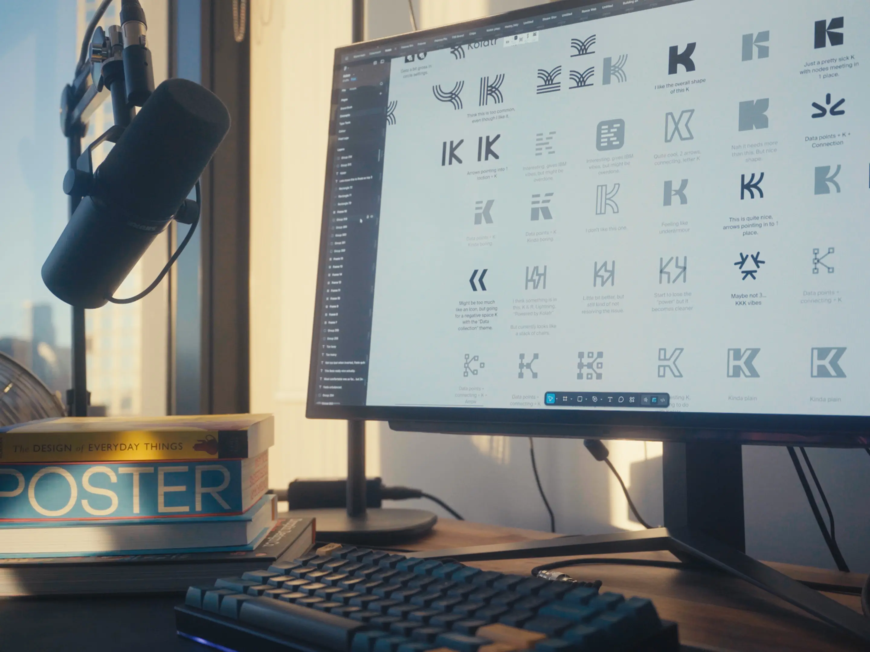Desk setup with a black microphone, stacked design books including 'Poster' and 'The Design of Everyday Things,' a keyboard, and a monitor displaying various logo designs.