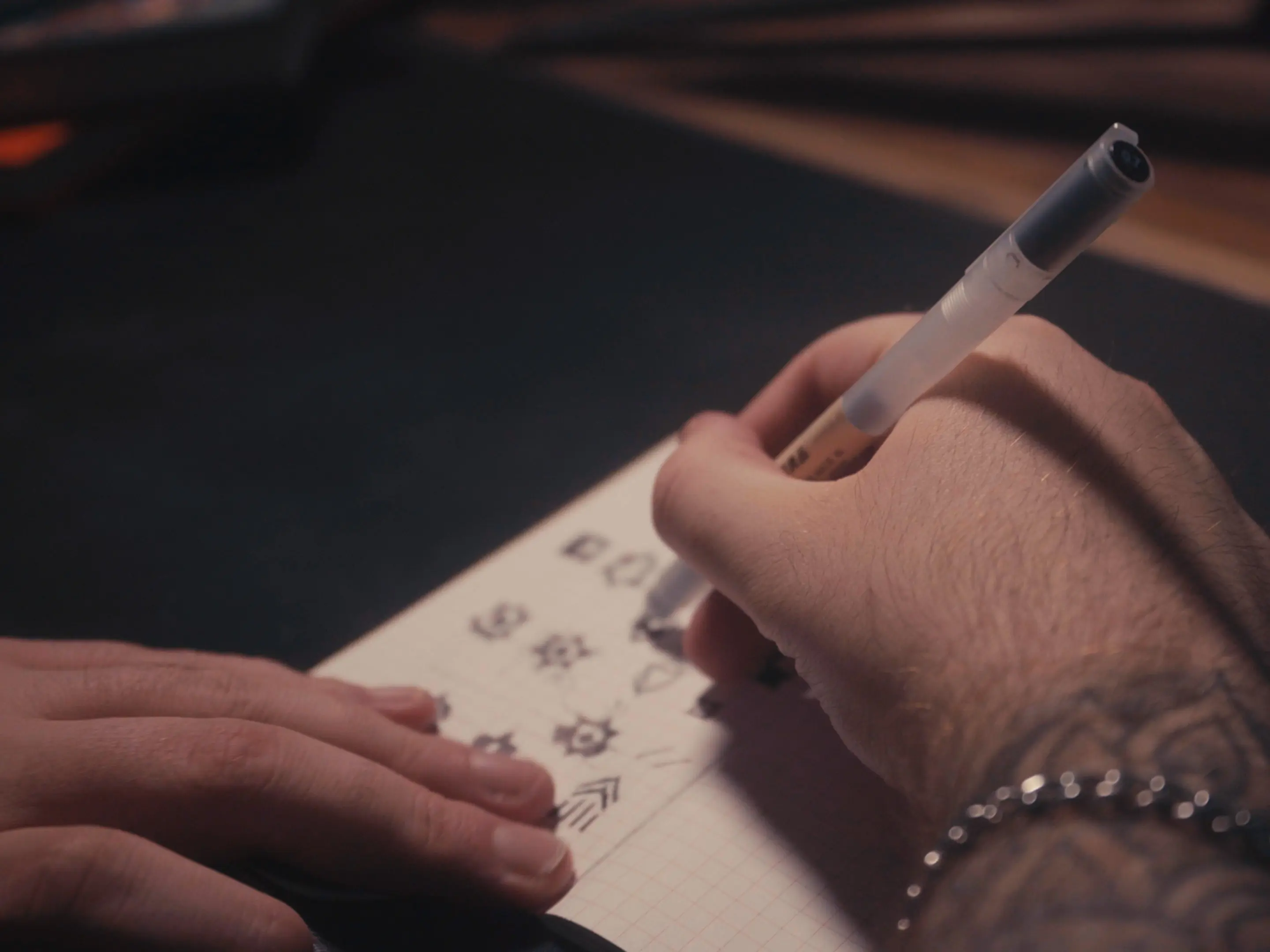 Person with tattooed wrist and bracelet drawing small icons in a notebook with a pen.