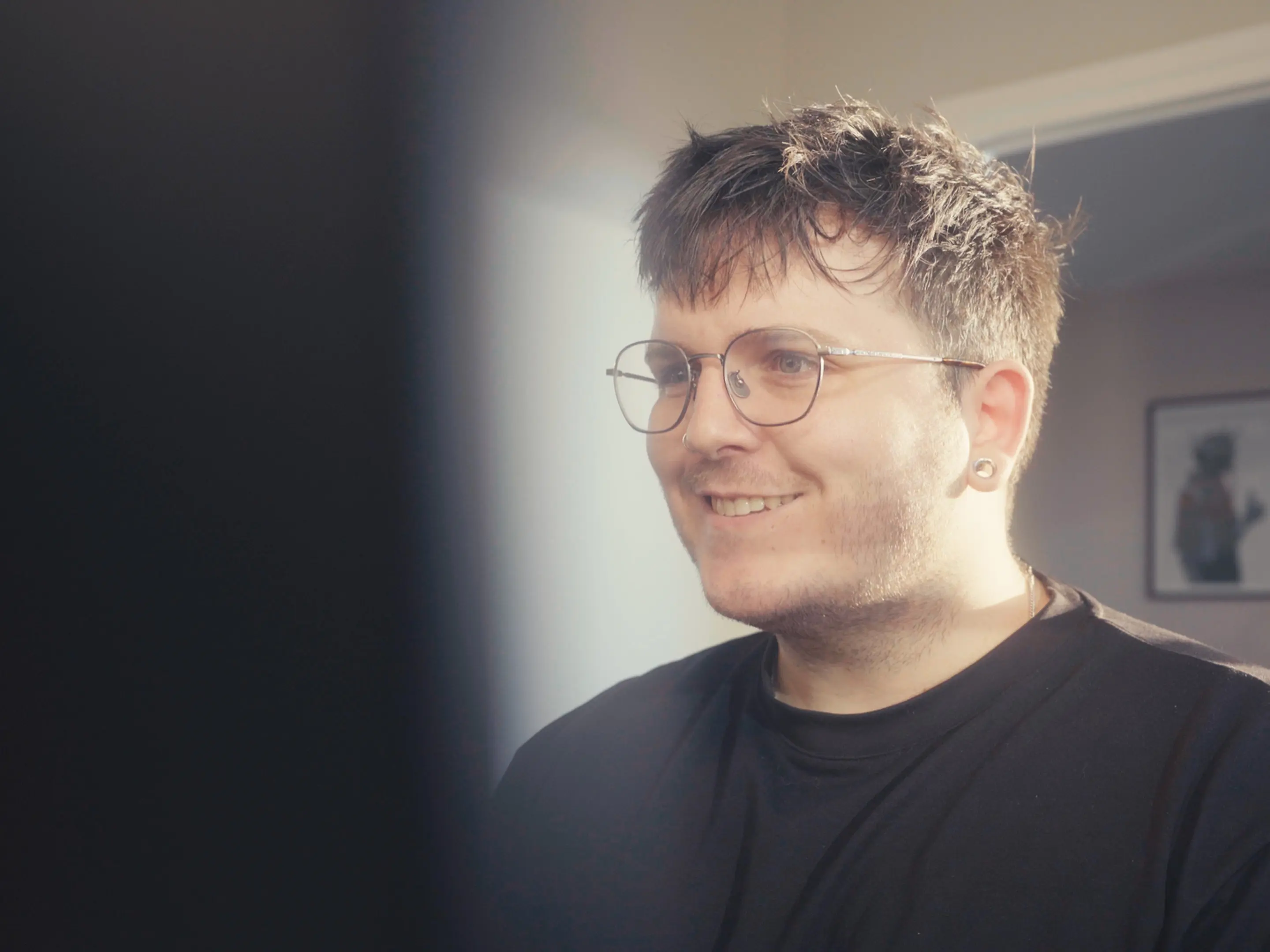 Smiling man with glasses and short messy hair looking to the side in a softly lit room.