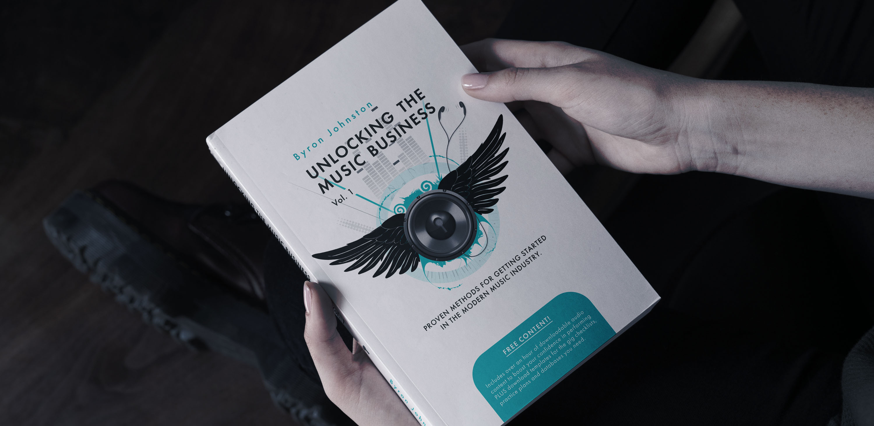 A woman’s hands holding a book in a dark room, the cover showing a digitally illustrated speaker with wings, surrounded by musical elements like a digitiser, headphones, and musical notes.