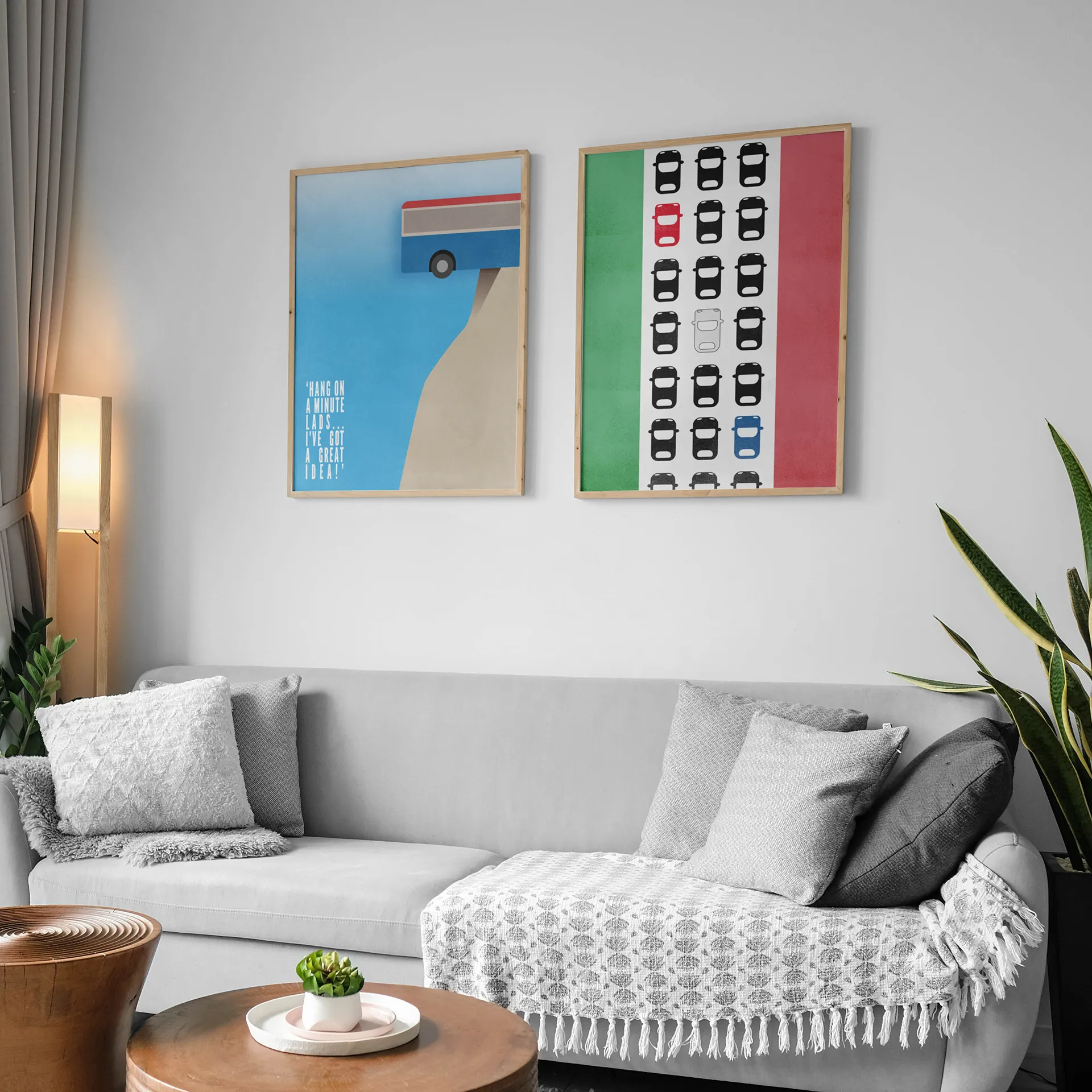 Two Framed minimalist posters above grey sofa. One shows a coach on the edge of a cliff over a blue sky; the other, a traffic jam of black cars with a red, blue, and white car on an Italian flag.