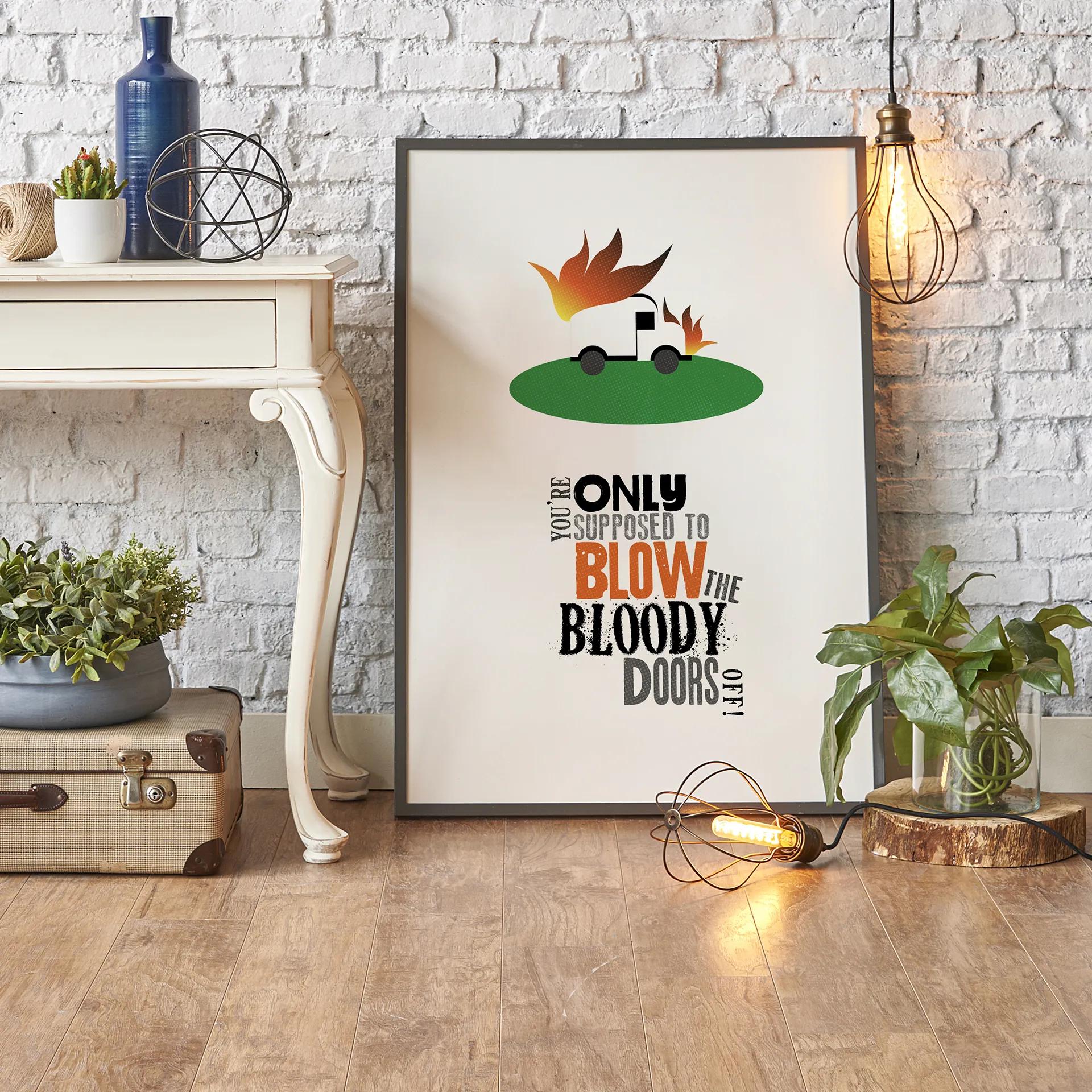 Framed poster next to a decorative side table and wire lights, showing a white van with orange flames on a green oval, with the quote ‘You’re only supposed to blow the bloody doors off’ below.