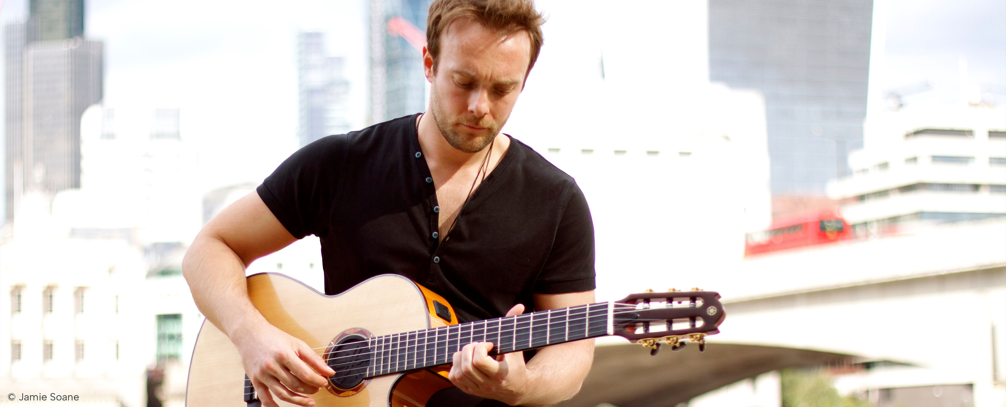 Photo of book author Byron Johnston sitting on a wall by the River Thames, playing a guitar. Photo by Jamie Soane.