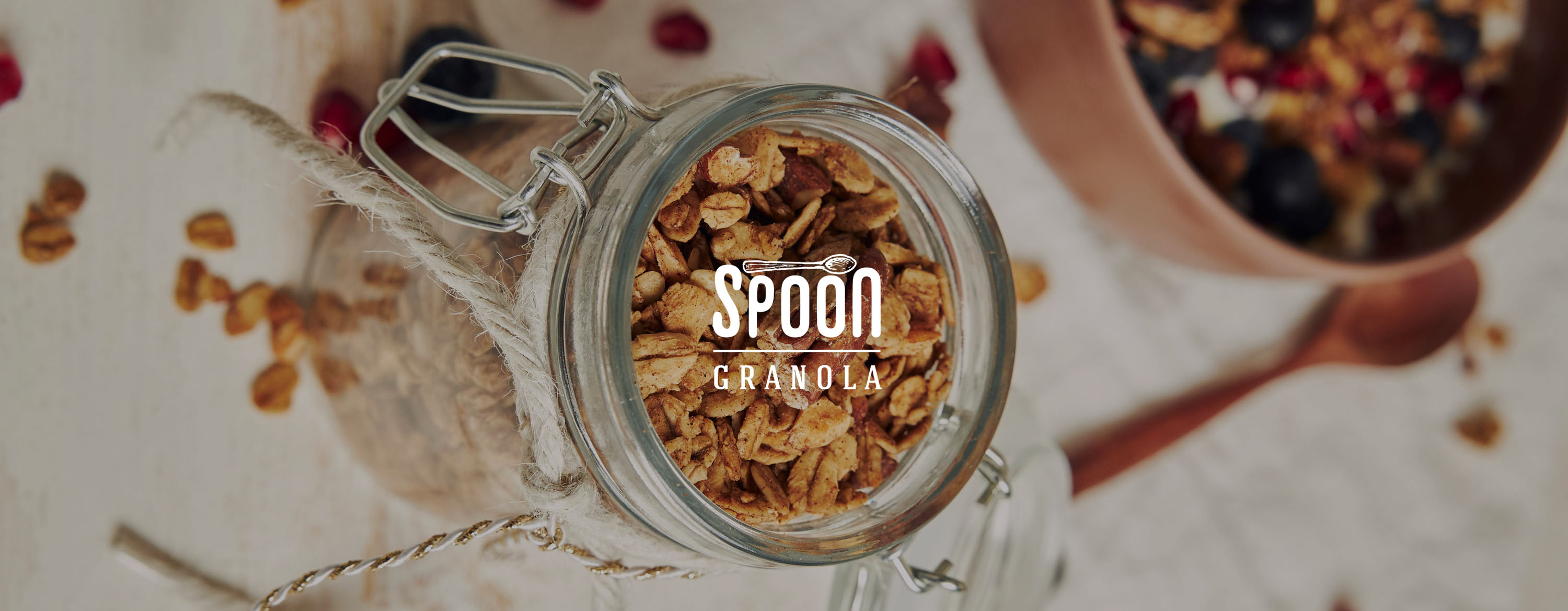 White hand-drawn Spoon Granola logo overlaid on an overhead photo of an open glass jar of granola.
