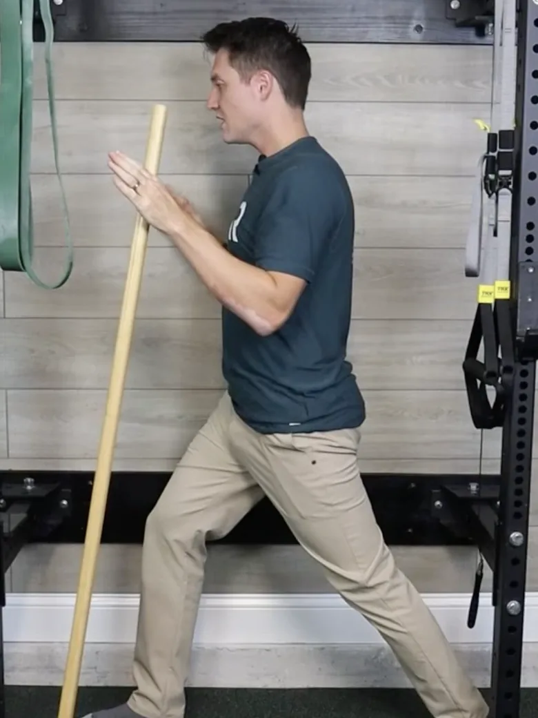 Dr. Michael Derry demonstrating a staggered stance balance hold, one leg stepped back, holding a balance stick for support