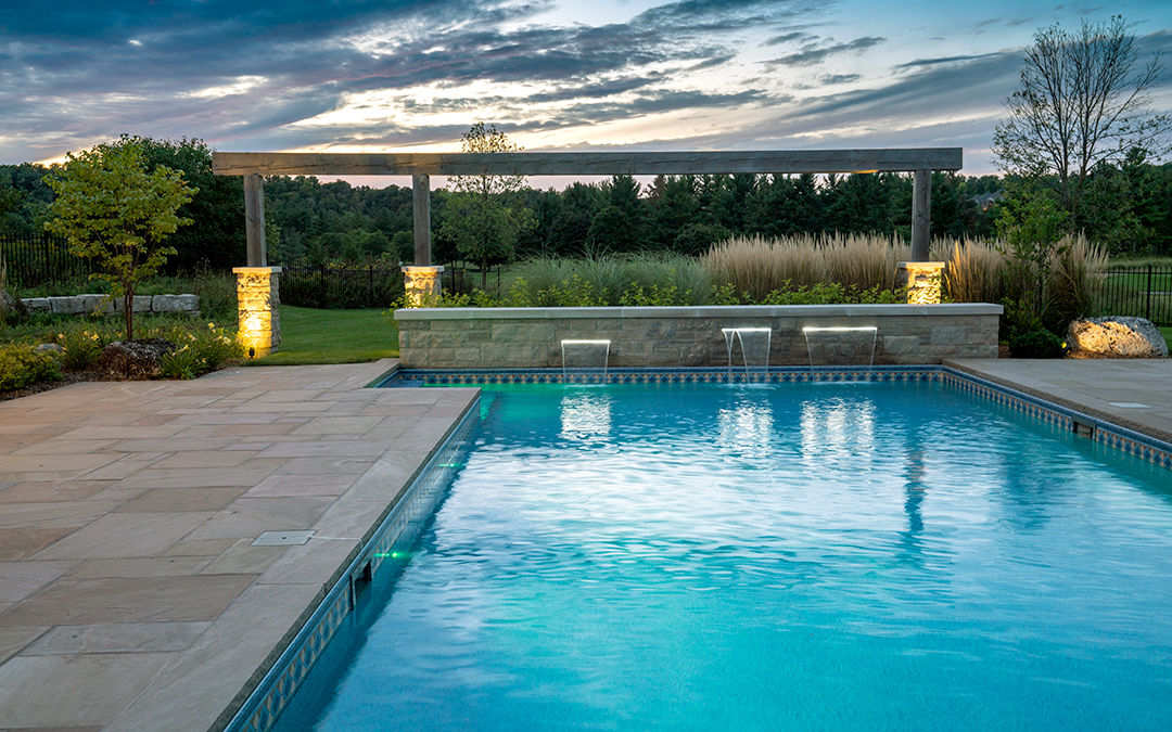 An outdoor pool with a large stone walkway and mini waterfall with landscape lighting and wooden arch.