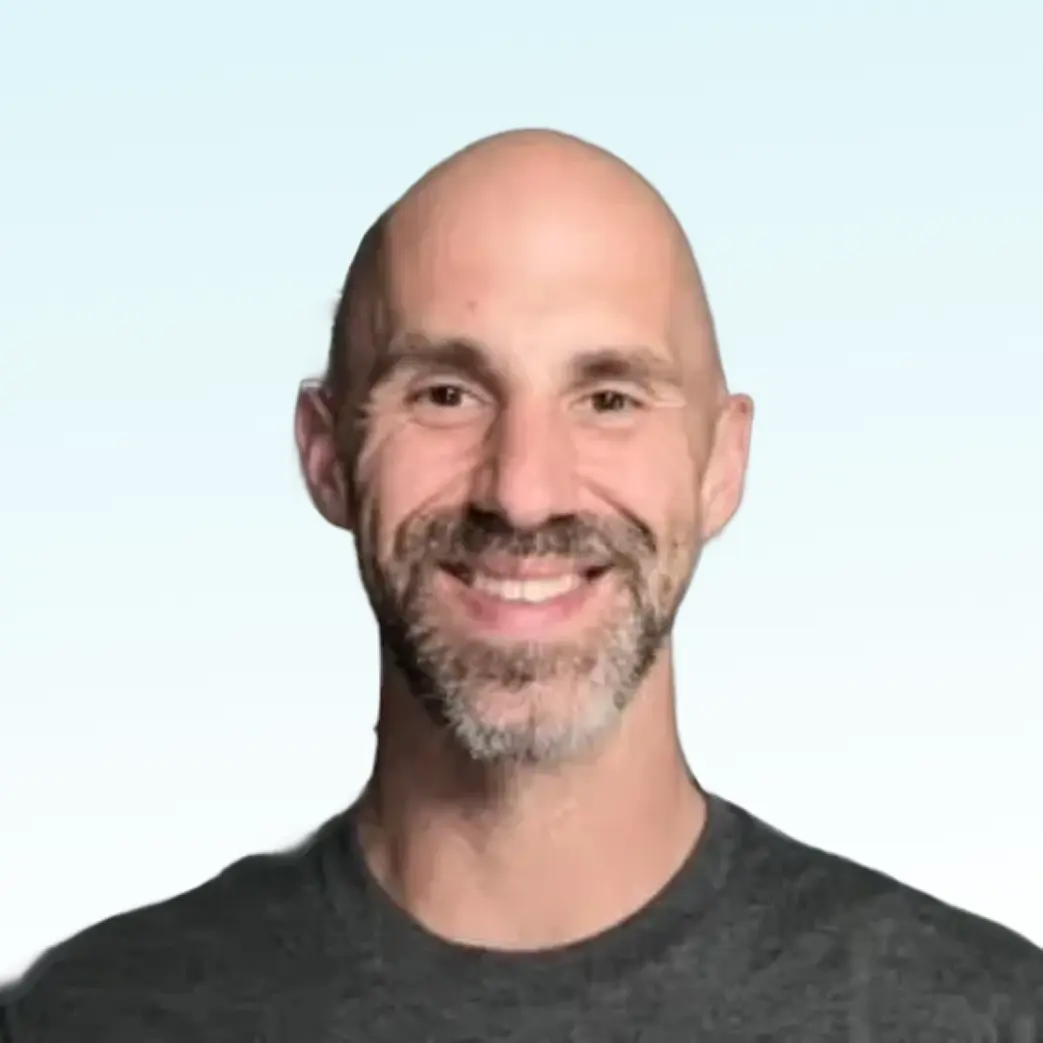 Smiling bald man with a trimmed beard wearing a dark gray shirt against a light blue background.