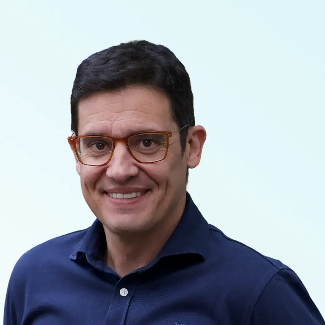 Smiling man with short dark hair and brown glasses wearing a navy blue shirt against a light background.