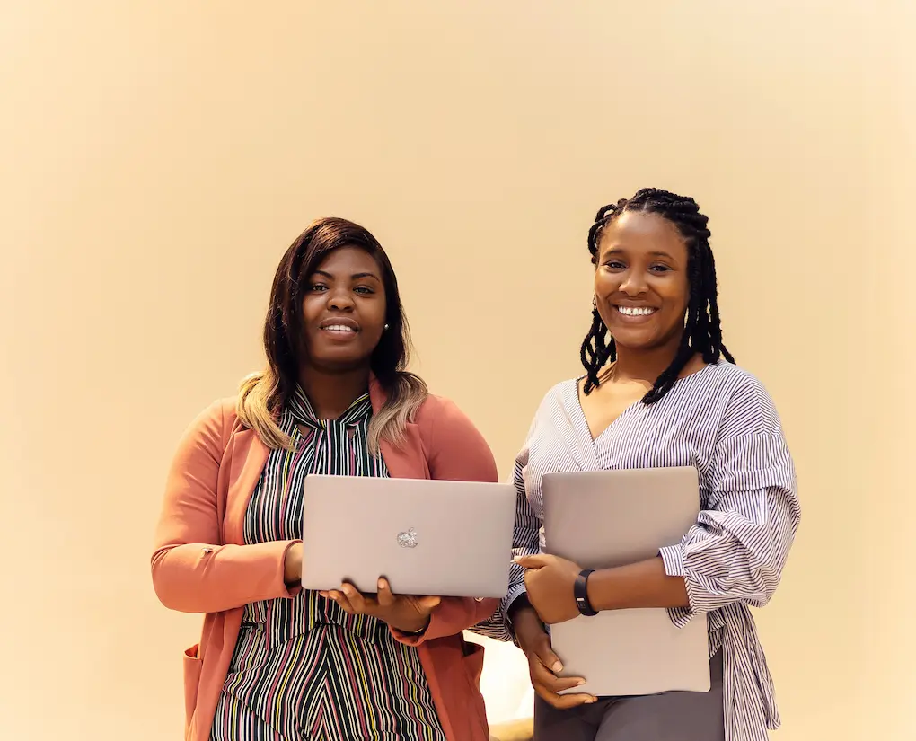 Two smiling women holding laptops standing against a plain beige background.