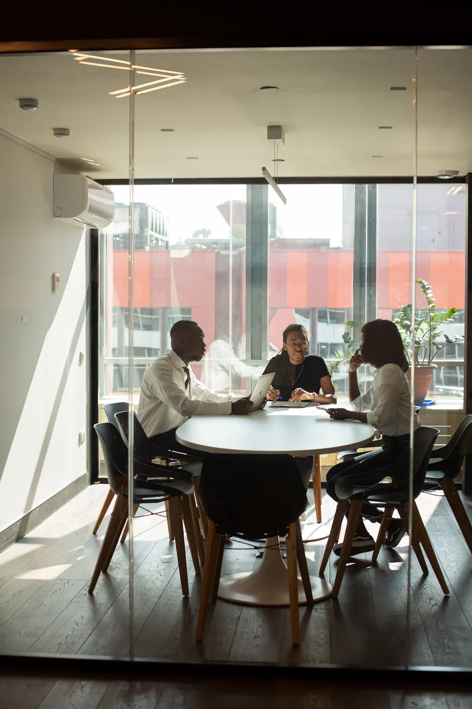 Three people sit at a round table in a sunlit office, discussing with a laptop and documents.