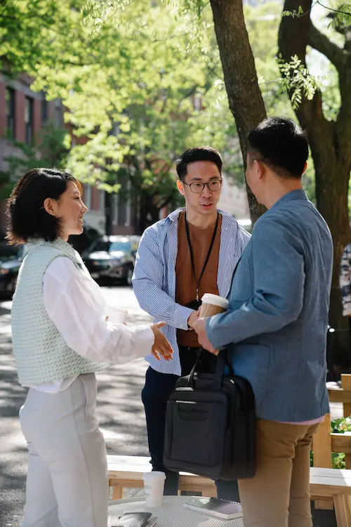 Three people having an outdoor conversation with coffee cups and a briefcase in a sunny park.