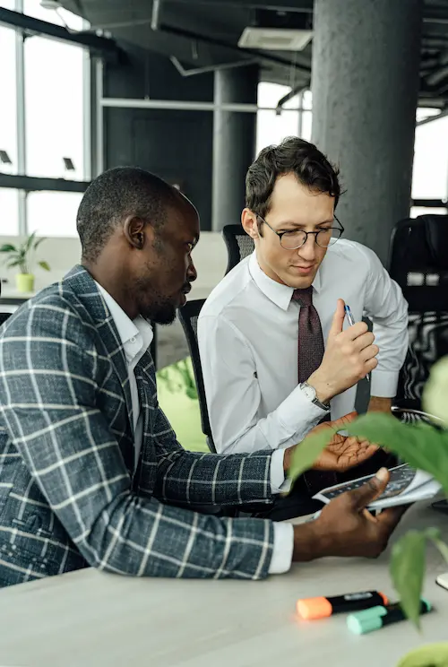 Two businessmen discuss a tablet while seated at an office desk with highlighters and plants nearby.