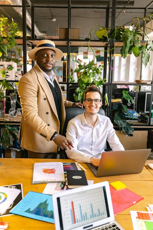 Two men smiling in an office with laptops, documents, and plants around a wooden desk.