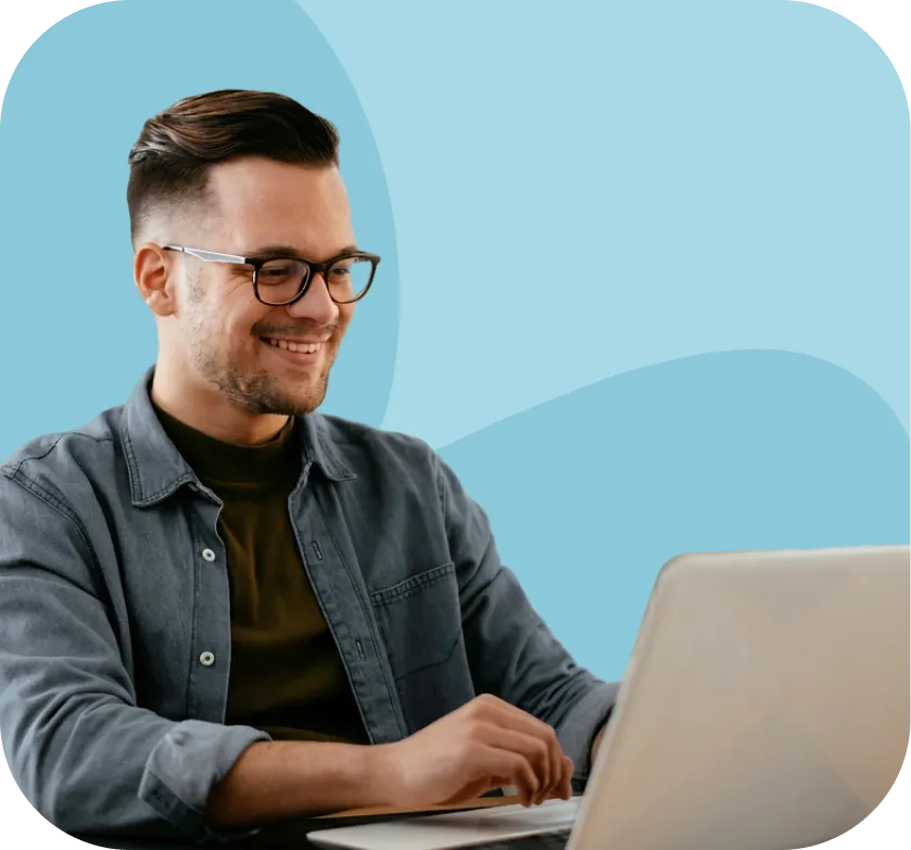 Smiling man with glasses and short hair wearing a dark shirt and denim jacket working on a laptop.
