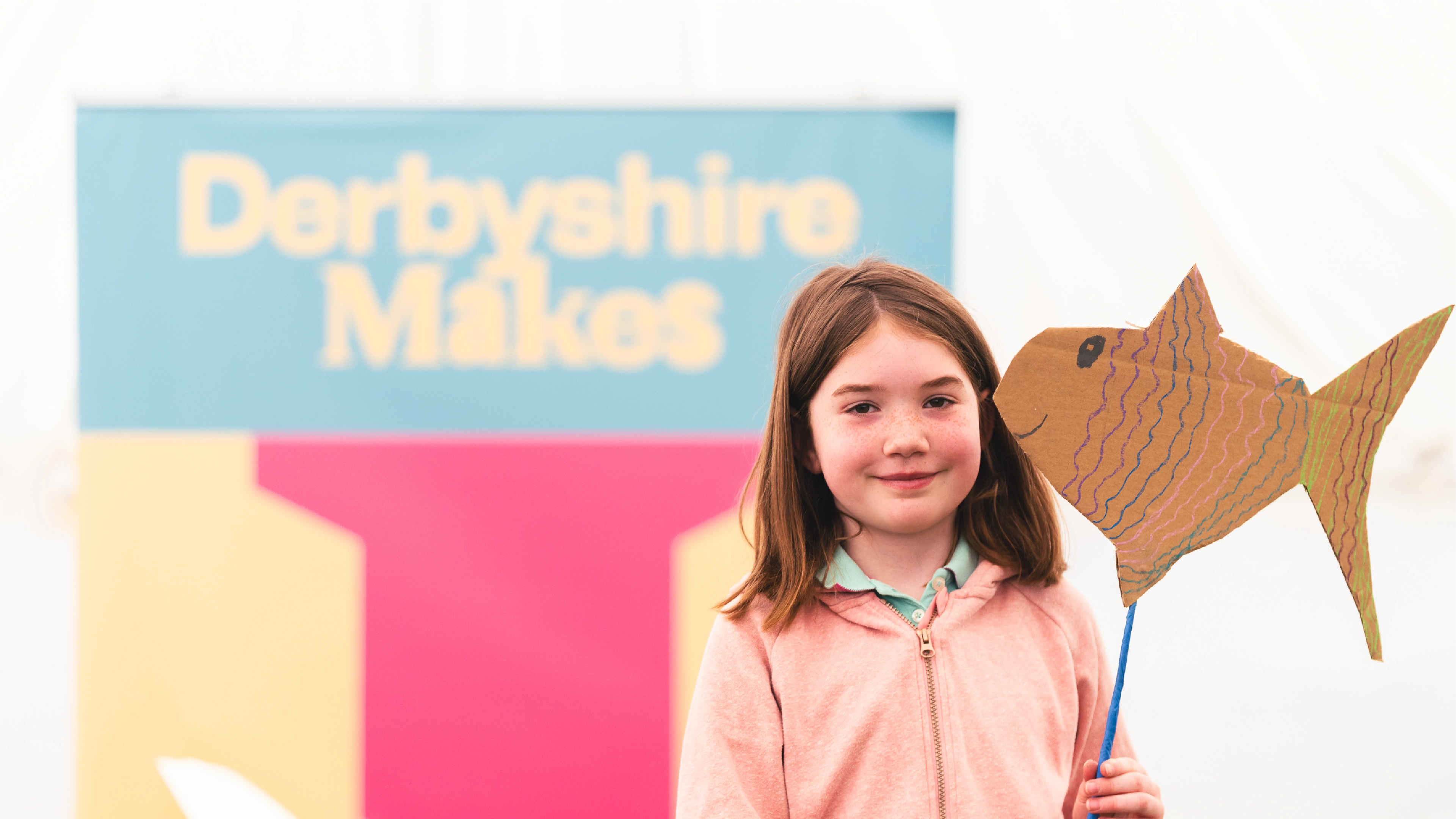Girl stood with her paper fish artwork at Derbyshire Makes festival in front of a banner with the branding created by Sanna Studio