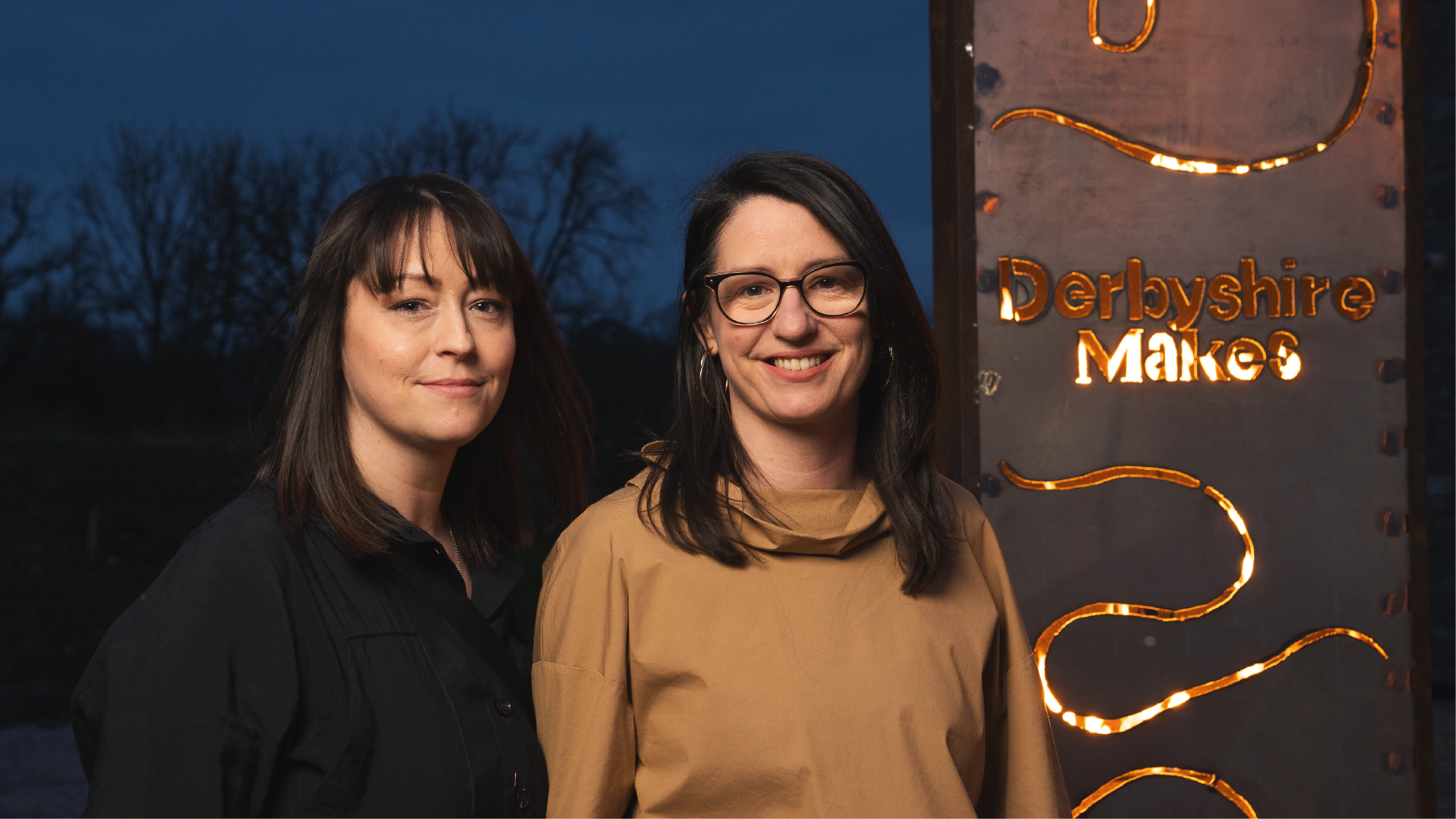 2 ladies stood next to a banner created from steel and fire with a design using the Derbyshire Makes brand identity created by Sanna Studio
