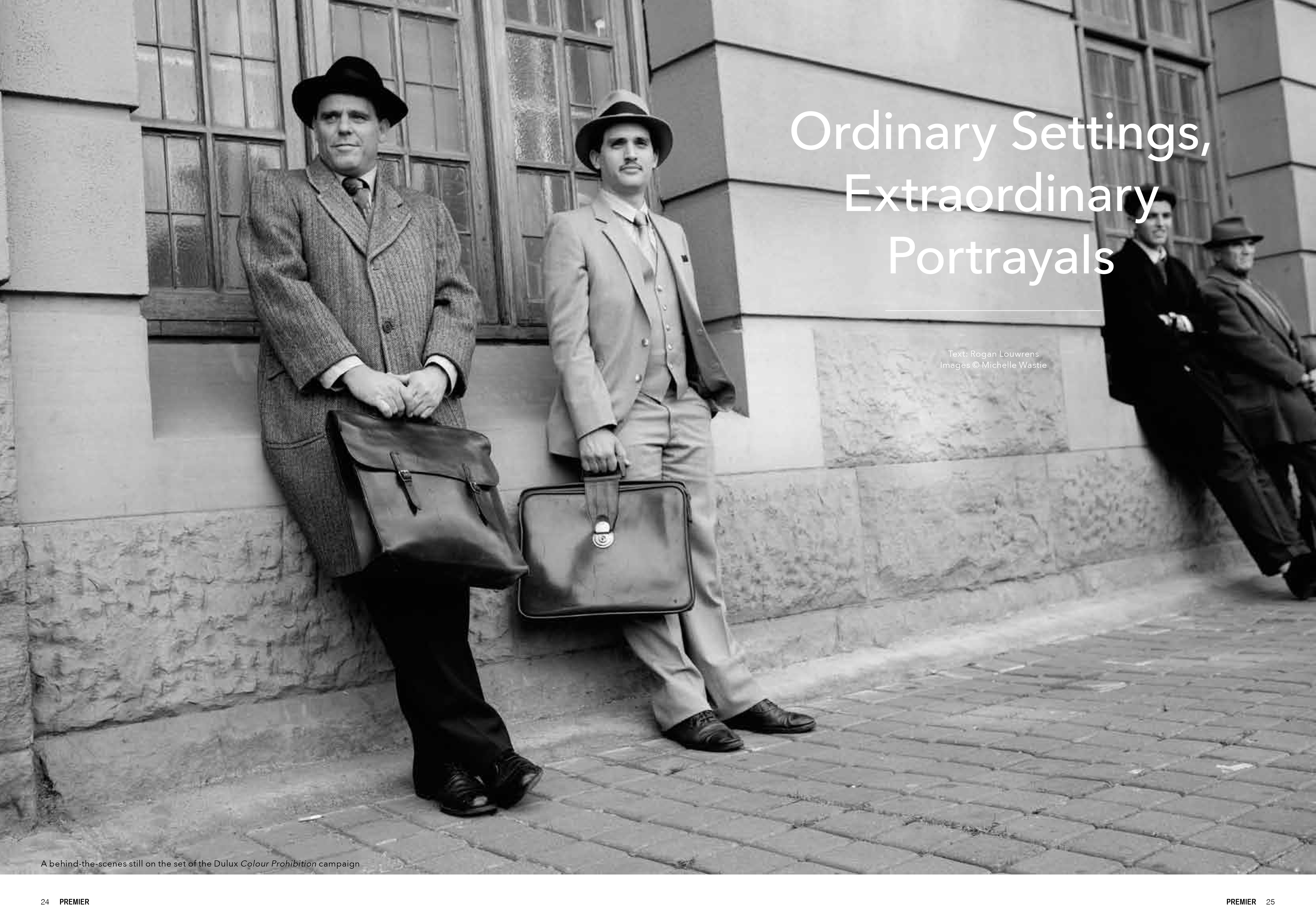Black-and-white photo of four men dressed in vintage suits and hats leaning against a stone building with briefcases.