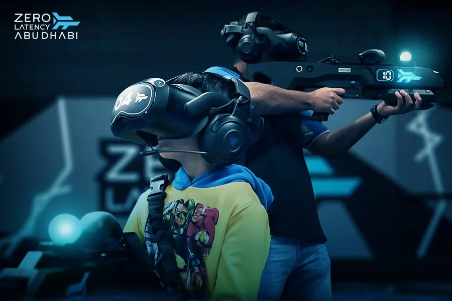 Player in a yellow shirt wearing a VR headset, with another player aiming a blaster behind