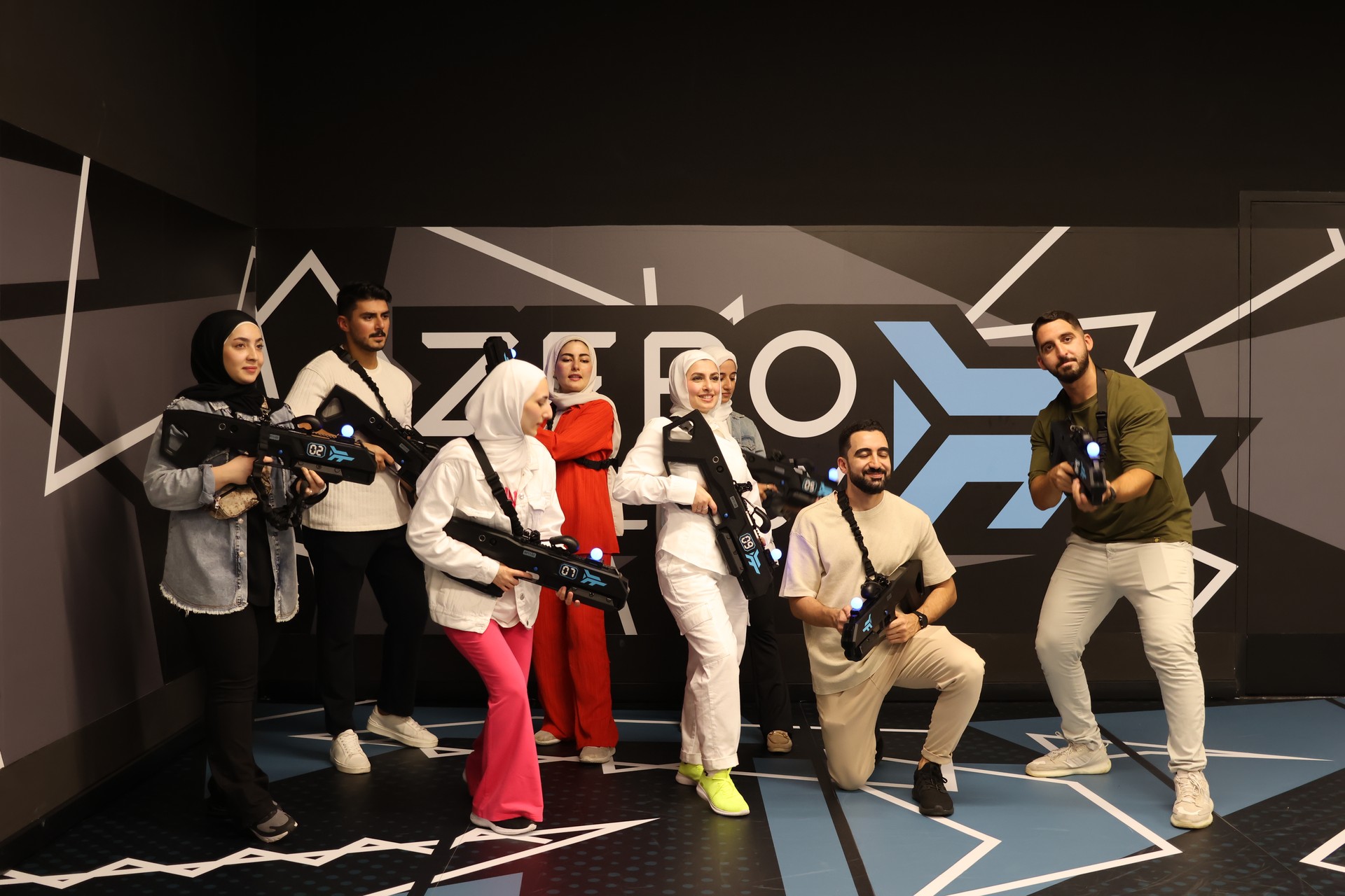 A group of eight people holding VR blasters pose together in front of a Zero Latency wall.