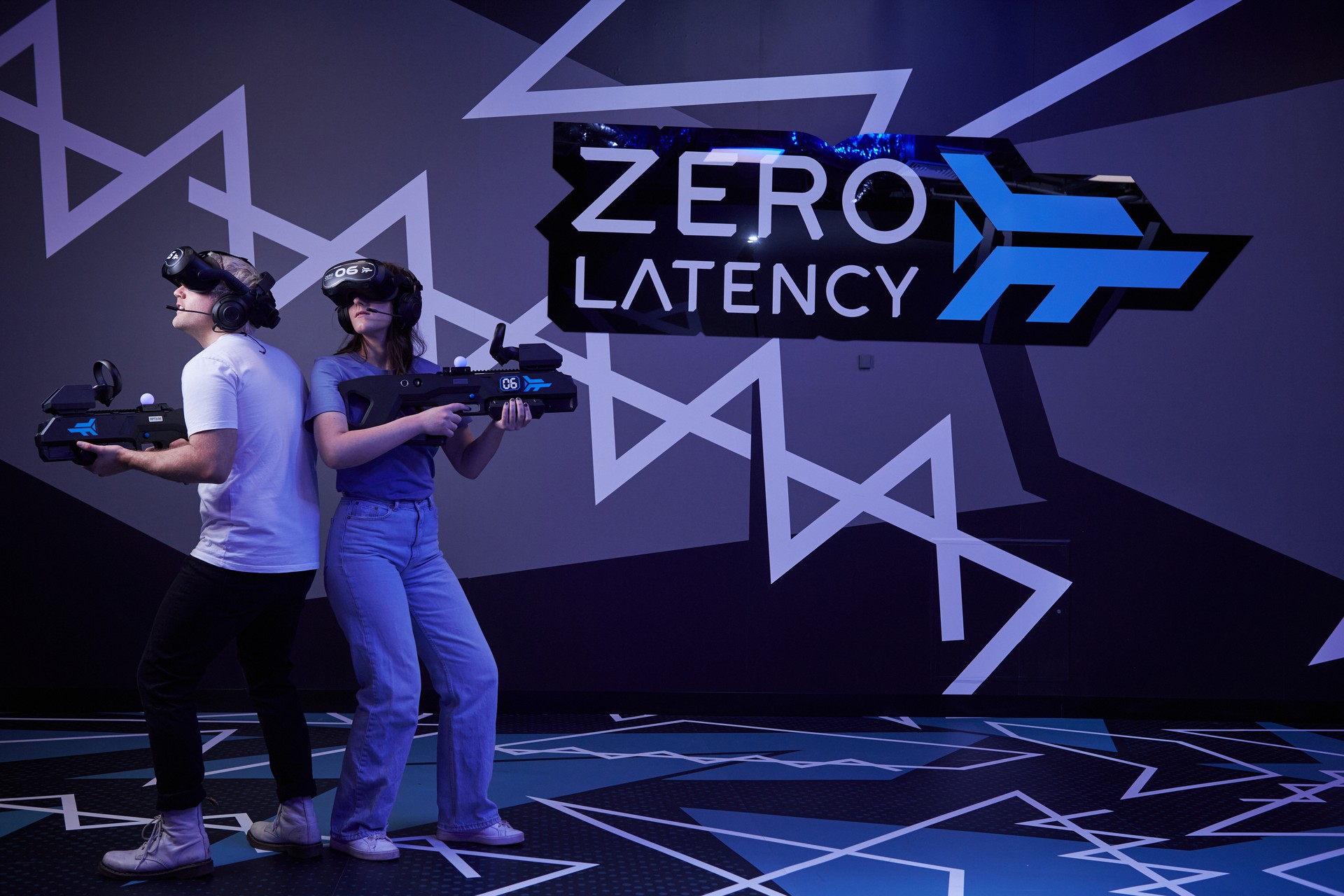 Two players in VR headsets stand back to back holding blasters beneath a Zero Latency sign.