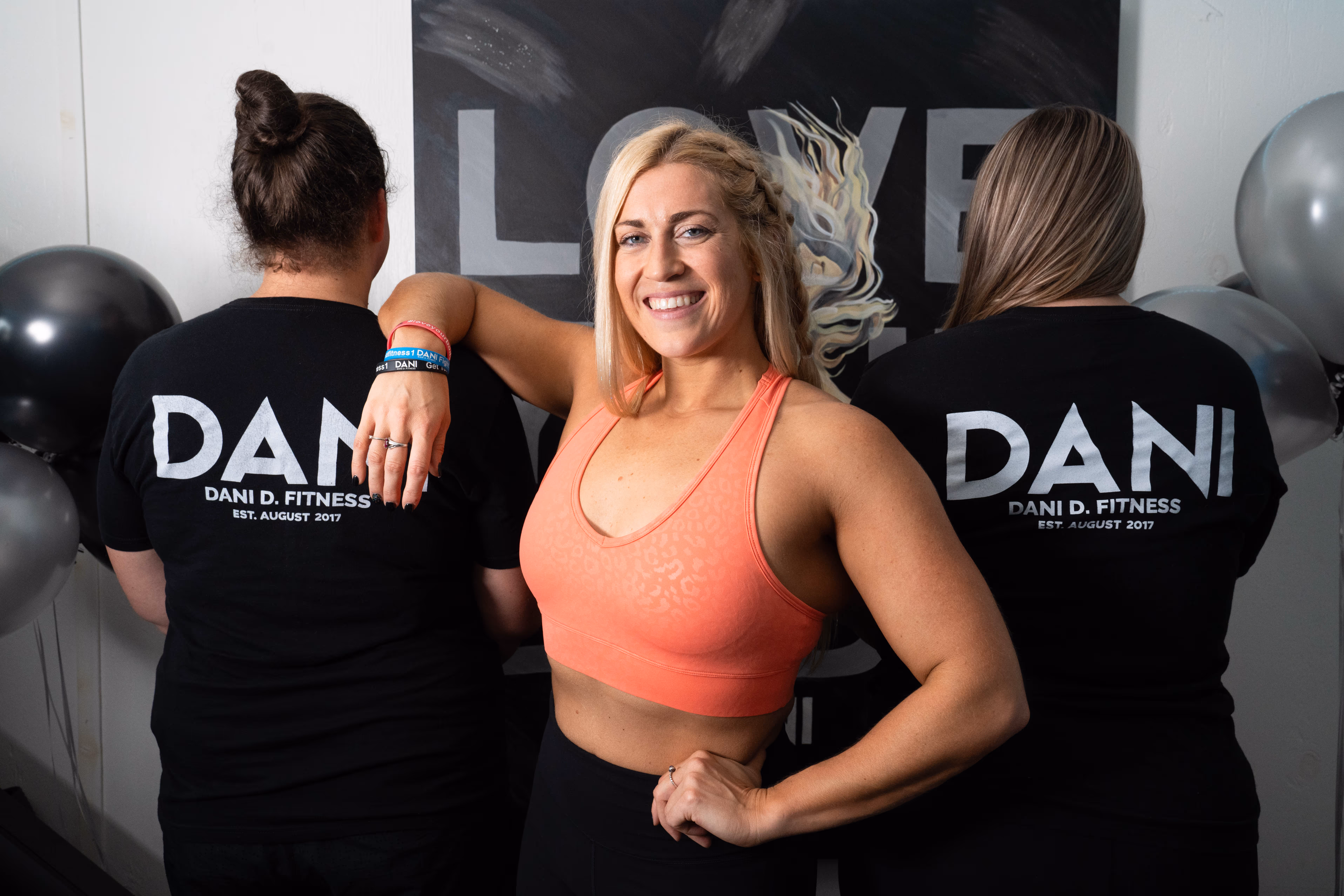 A woman in an orange sports bra smiles in front of two women wearing "Dani D. Fitness" T-shirts. Black balloons are in the background.