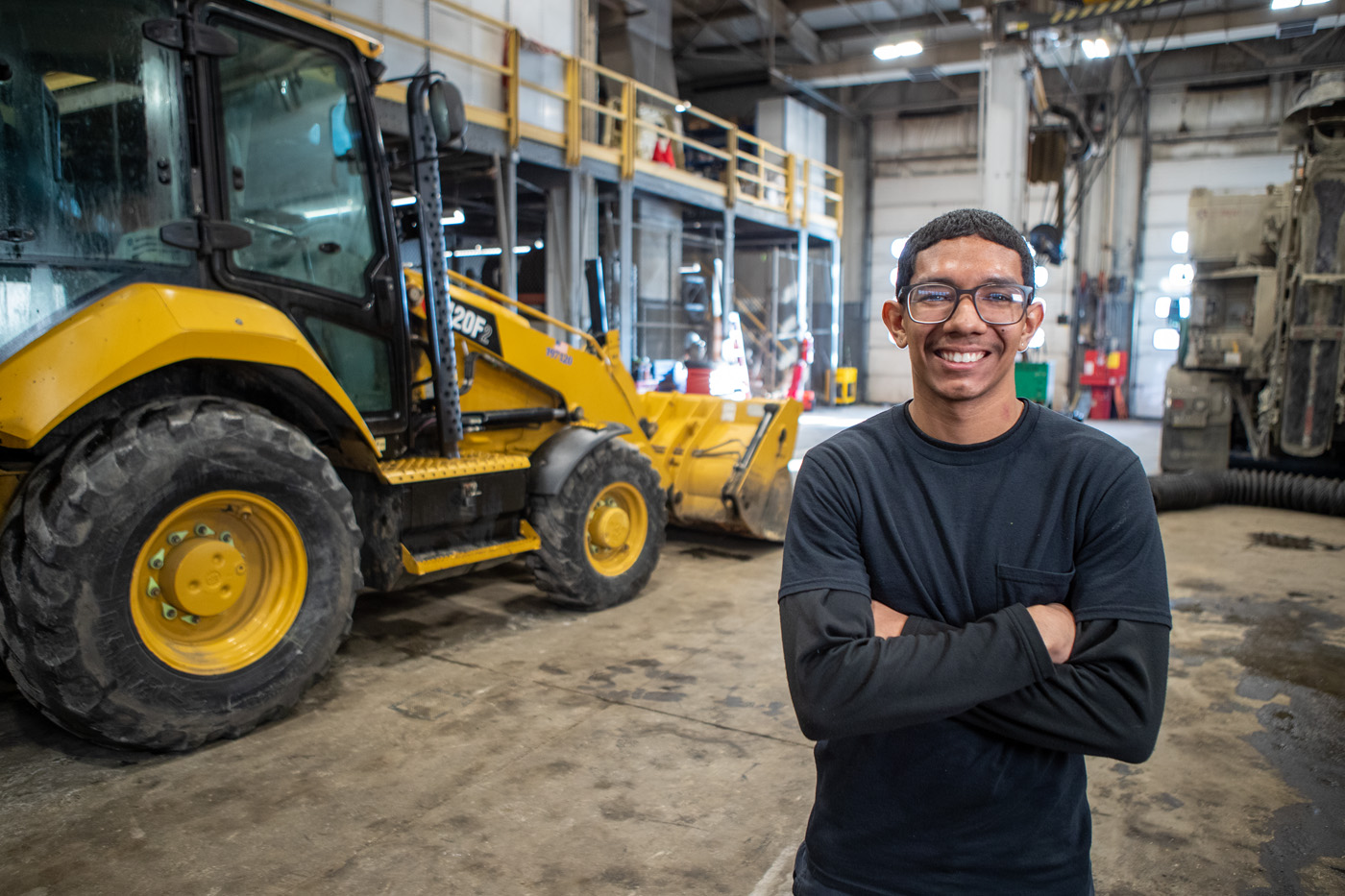 Auto student and mentor in front of an excavator in the garage during an I AM SDP commercial. 