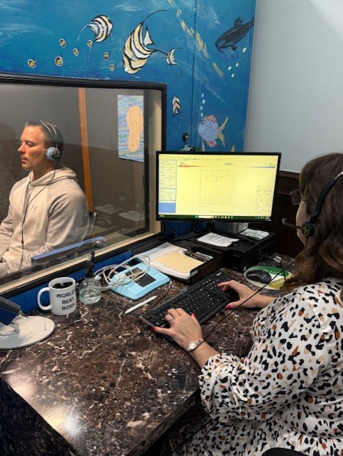 Female audiologist wearing a white coat and face shield fitting a hearing aid for a seated client with blond hair tied back with a decorative hair clip.