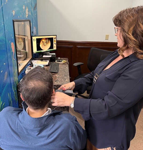Audiologist wearing a white coat and face shield fitting a hearing aid on a seated patient in a bright office with a green wall and anatomical ear diagrams.