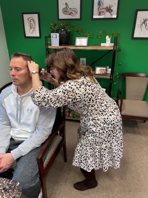Audiologist wearing a white coat and clear face shield fitting a hearing aid on a seated patient.