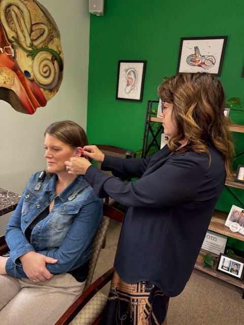 Audiologist fitting a hearing aid on a seated woman with hair in a bun and a decorative hair clip.