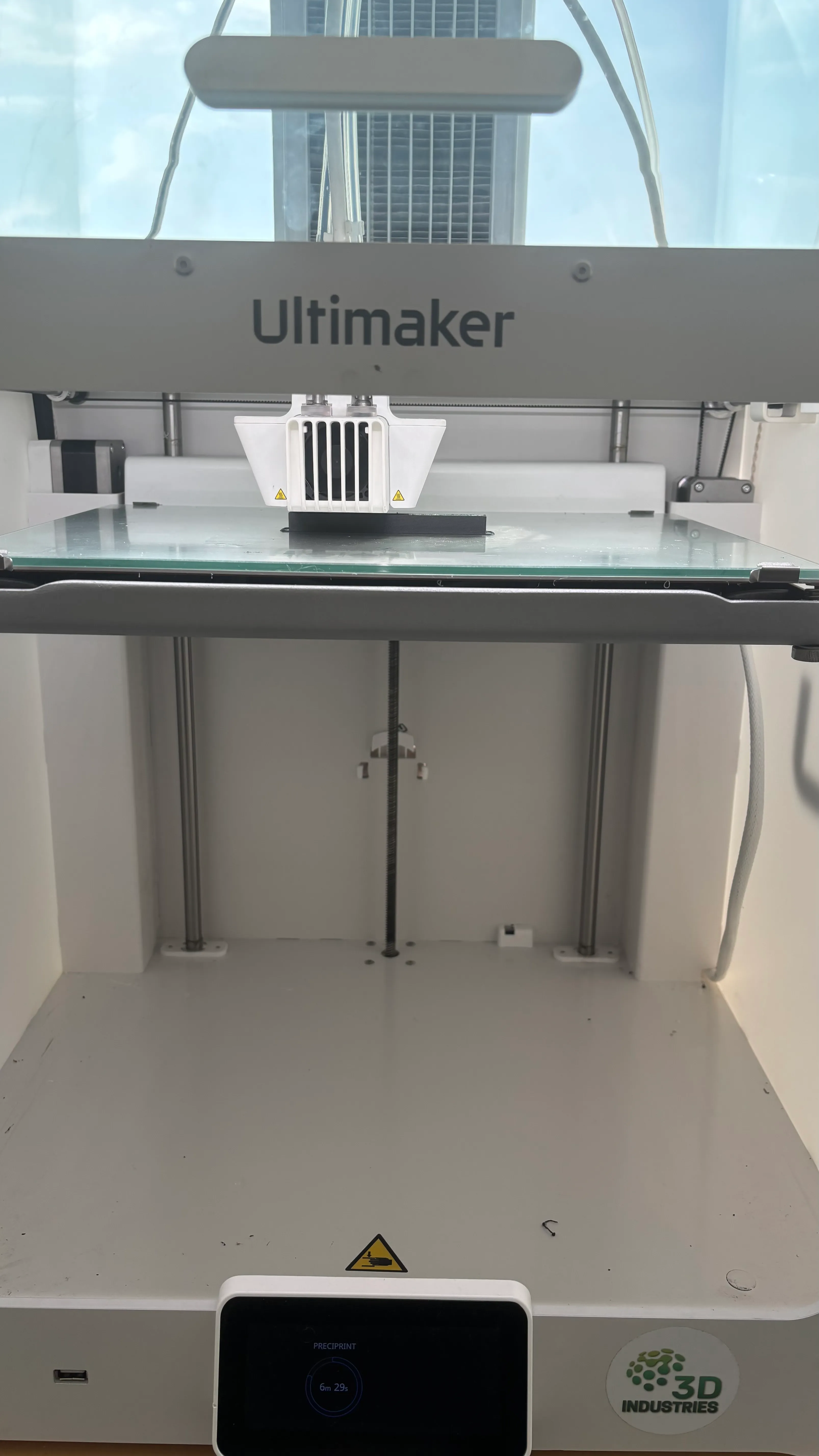 Imprimante 3D Ultimaker vide sur une table en bois avec une fenêtre et une voiture bleue en arrière-plan.