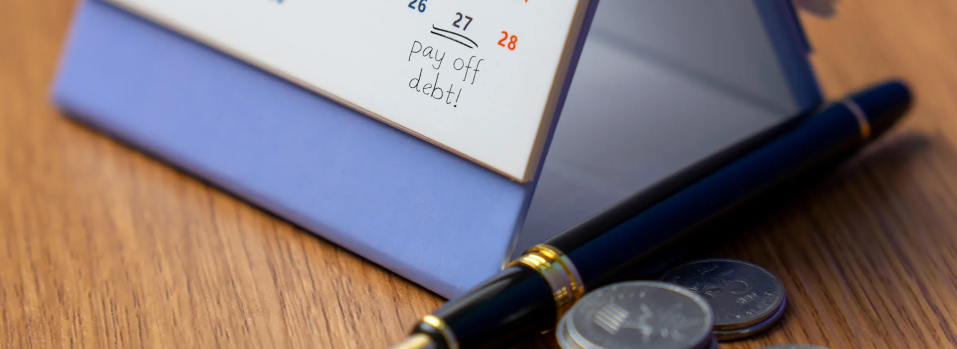 A desk calendar with "pay off debt!" marked on a date beside a pen and a few stacks of coins