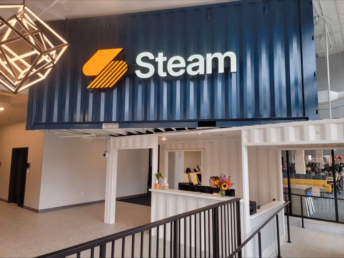A modern office lobby featuring a reception desk housed in a repurposed white shipping container. Above, a dark blue container displays an illuminated ‘Steam’ logo in white and orange.