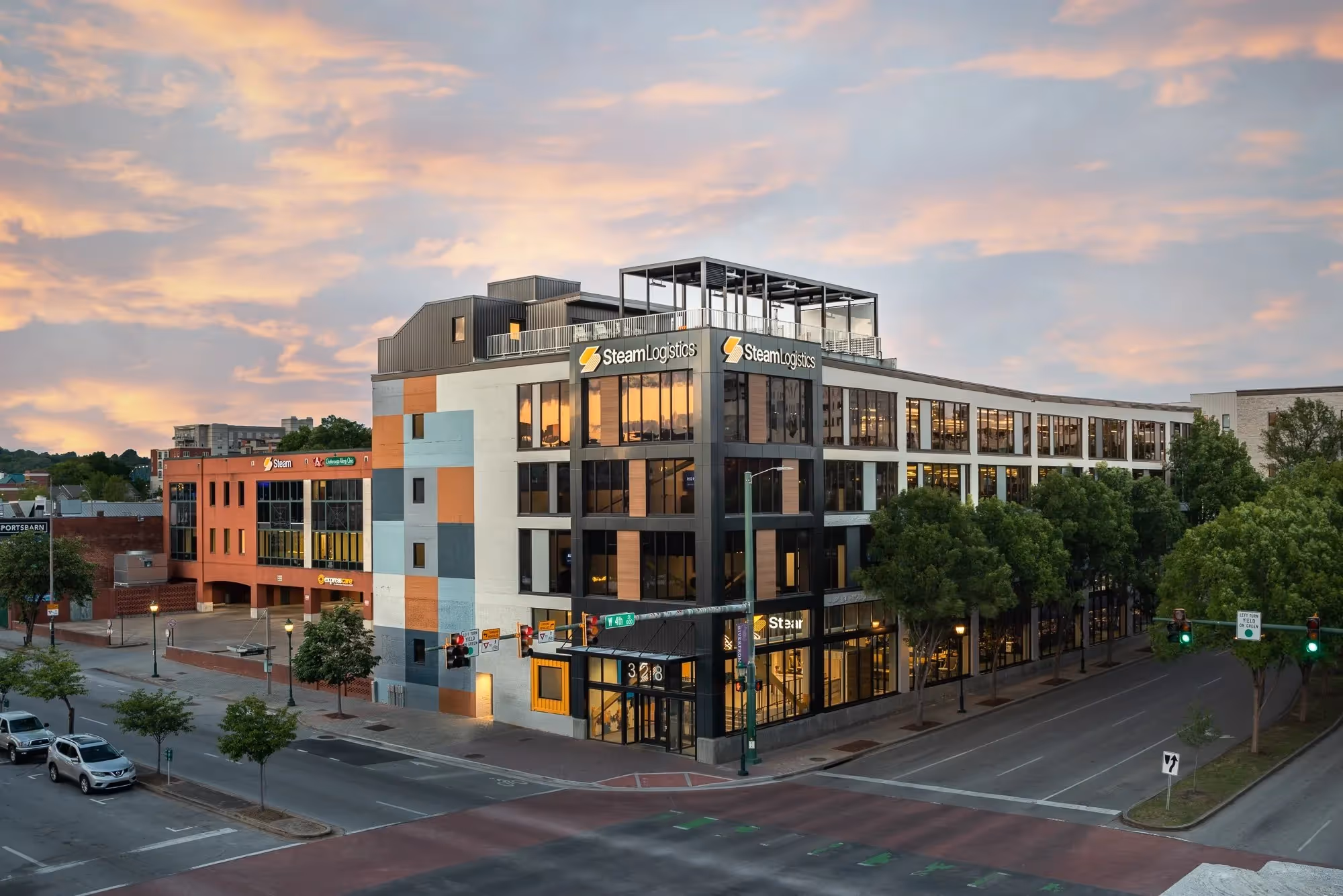 A modern, multi-story building with large windows and the Steam Logistics logo, set against a sunset sky in an urban setting.