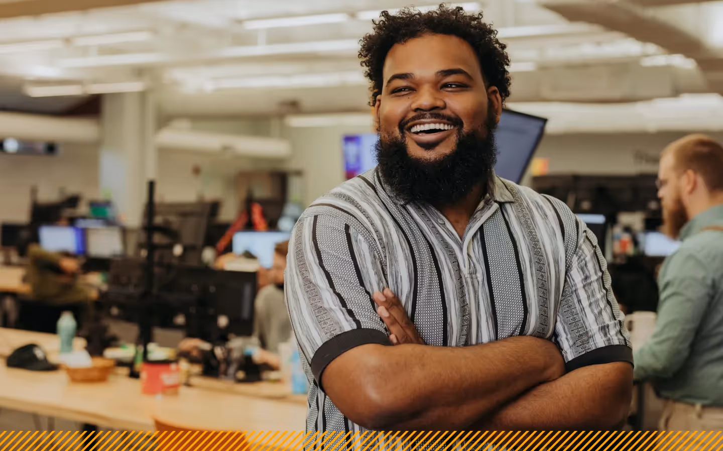 A bearded man in a striped shirt smiling with arms crossed in an open-plan office environment with employees working at desks in the background.