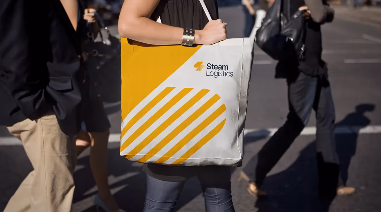 A person carrying a white tote bag with yellow diagonal stripes and the Steam Logistics logo while walking on a city street.