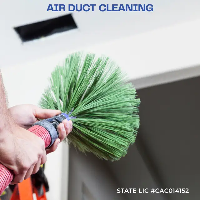 Hands holding a green brush cleaning an air duct vent in a white ceiling.