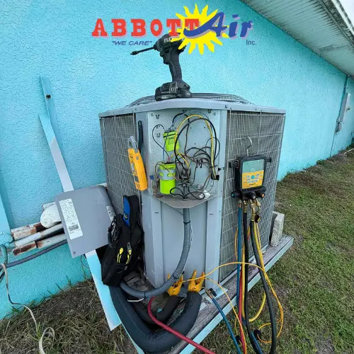 Outdoor HVAC unit with diagnostic tools and wiring, mounted next to a turquoise textured wall.