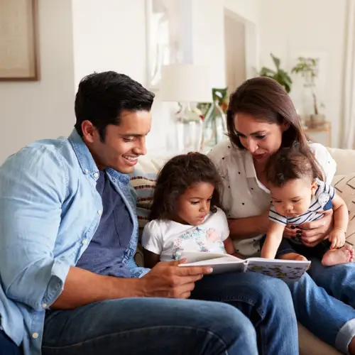 Parents sitting on a couch reading a book to their two young children in air conditioned room.