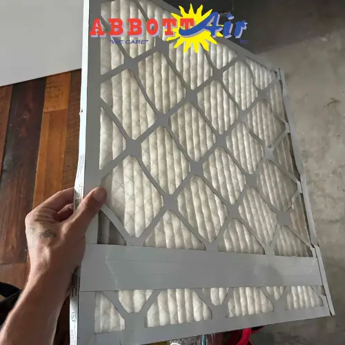 Hand holding a white rectangular air filter with diamond-patterned grid on one side and pleated filter material visible inside, against a background of wood floor and concrete wall, with a colorful Abbott Air logo in the top left corner.