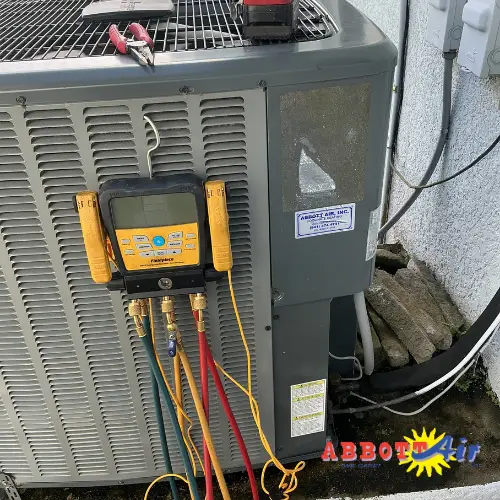 Gray outdoor air conditioning unit with a digital manifold gauge and connected colored hoses for HVAC measurements, situated next to a wall with electrical components.