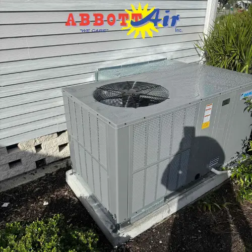 Outdoor HVAC air conditioning unit installed on a concrete slab next to a white wooden house with plants around it and a shadow cast on the unit.