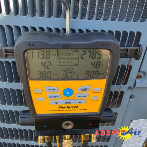 Fieldpiece digital HVAC manifold gauge attached to an air conditioning unit, displaying low and high pressure readings, temperature, and other system parameters.
