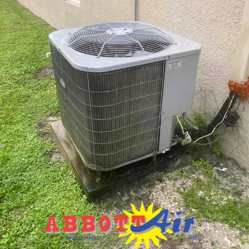 Outdoor HVAC air conditioning unit installed on a concrete pad beside a beige wall, surrounded by grass and some gravel, with an Abbott Air logo at the bottom.
