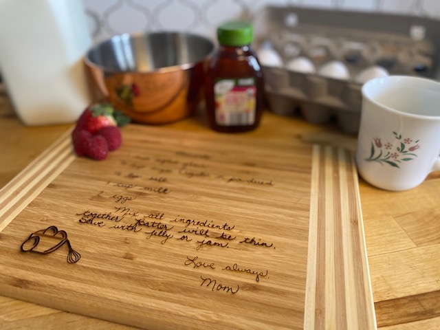 A standard size bamboo cutting board with personalized laser engraved family recipe by Forged One Two  in a kitchen with strawberries, eggs, honey, coffee cup and various other baking items.