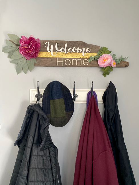 A reclaimed locally sourced rustic  wood  sign made by Forged One Two with large peonies, a painted gold metallic streak down the middle and 3D laser cut letters that read Welcome Home hanging over a coat rack in an entry way.