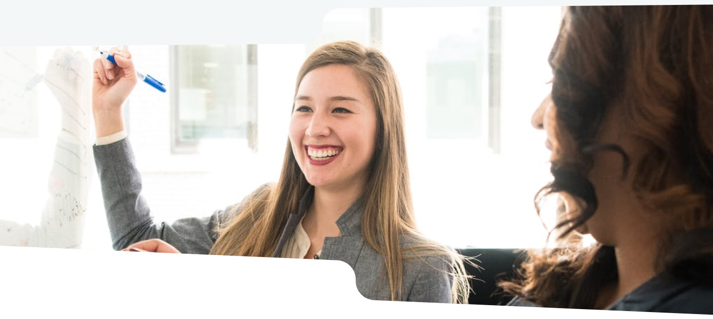 Photo of two women in an office writing on a whiteboard with a marker.