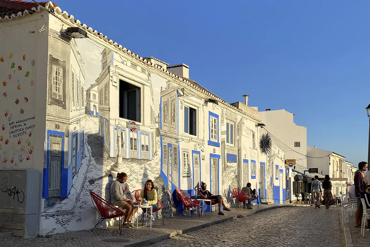 Street in Ericeira’s village centre with illustrated building façades and people sitting outside in the late-afternoon sun — a charming setting near many boutique hotels in Ericeira.