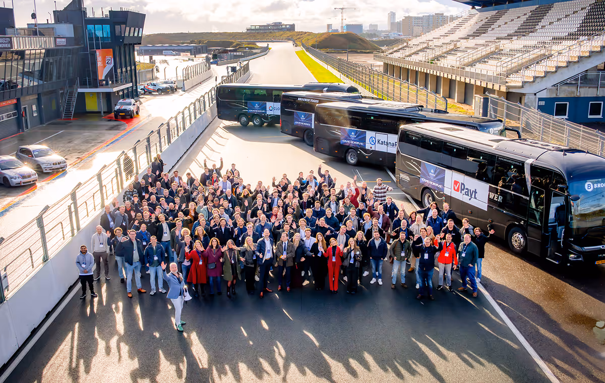 Group photo at Alumio Overdrive 2025 on a racetrack near buses with company logos, waving and smiling