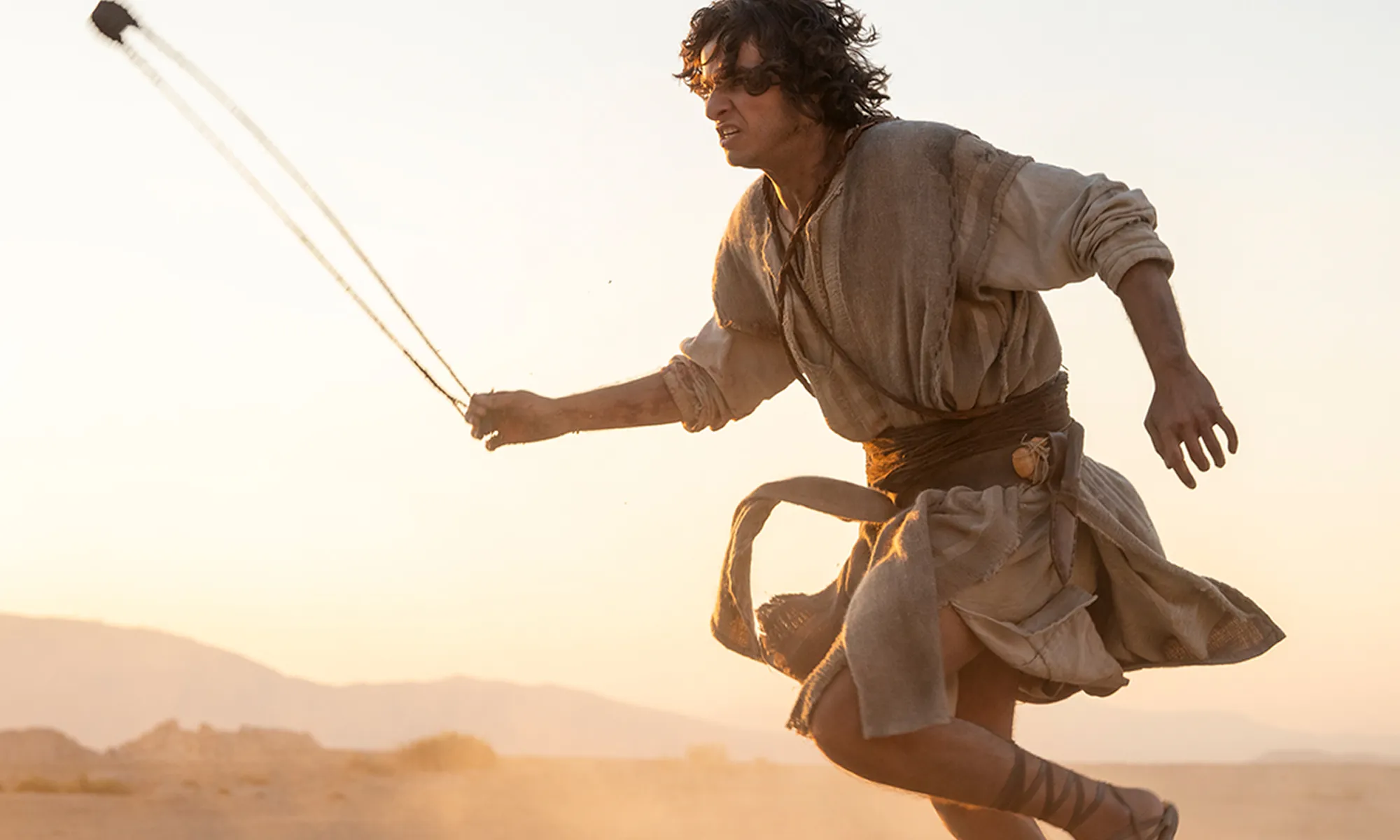 Man in ancient-style clothing wielding slingshot in a desert landscape with mountains in the background at sunset.